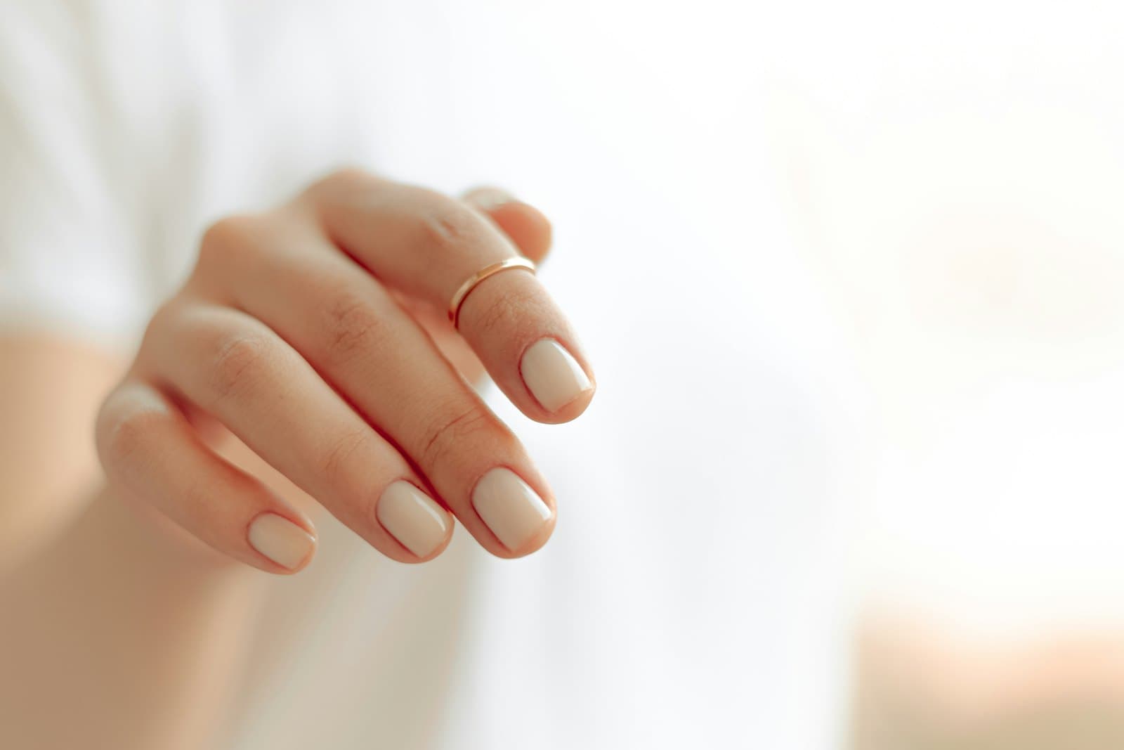 Close-up of a nude manicure and a thin gold ring against a soft white backdrop, editorial style